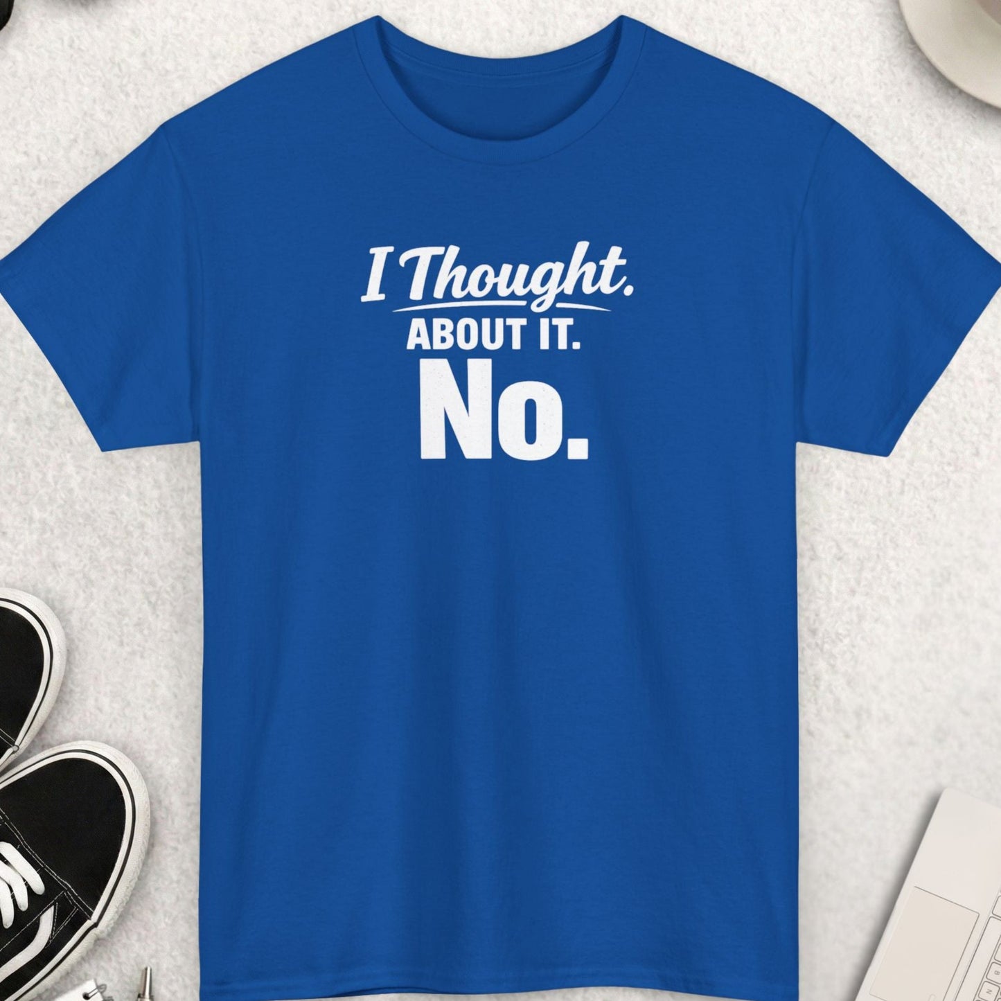 Blue t-shirt with 'I Thought About It. No.' text on a light gray surface with shoes, sunglasses, and a cup.