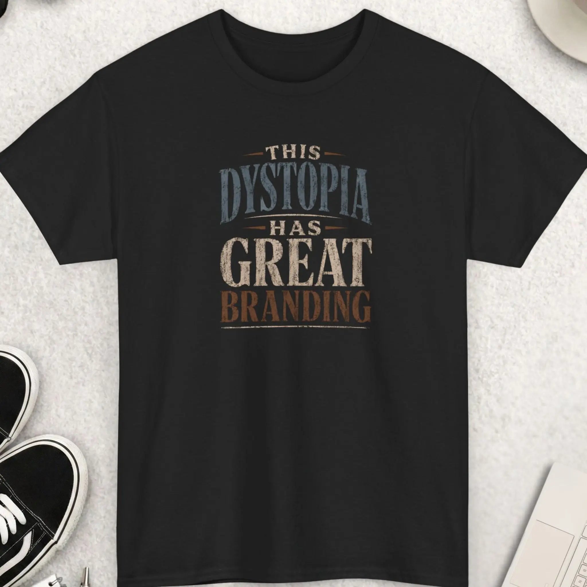 Distressed vintage typography t-shirt reading “This Dystopia Has Great Branding” in cream, steel blue, and burnt copper lettering on a black shirt with dark humor streetwear aesthetic.