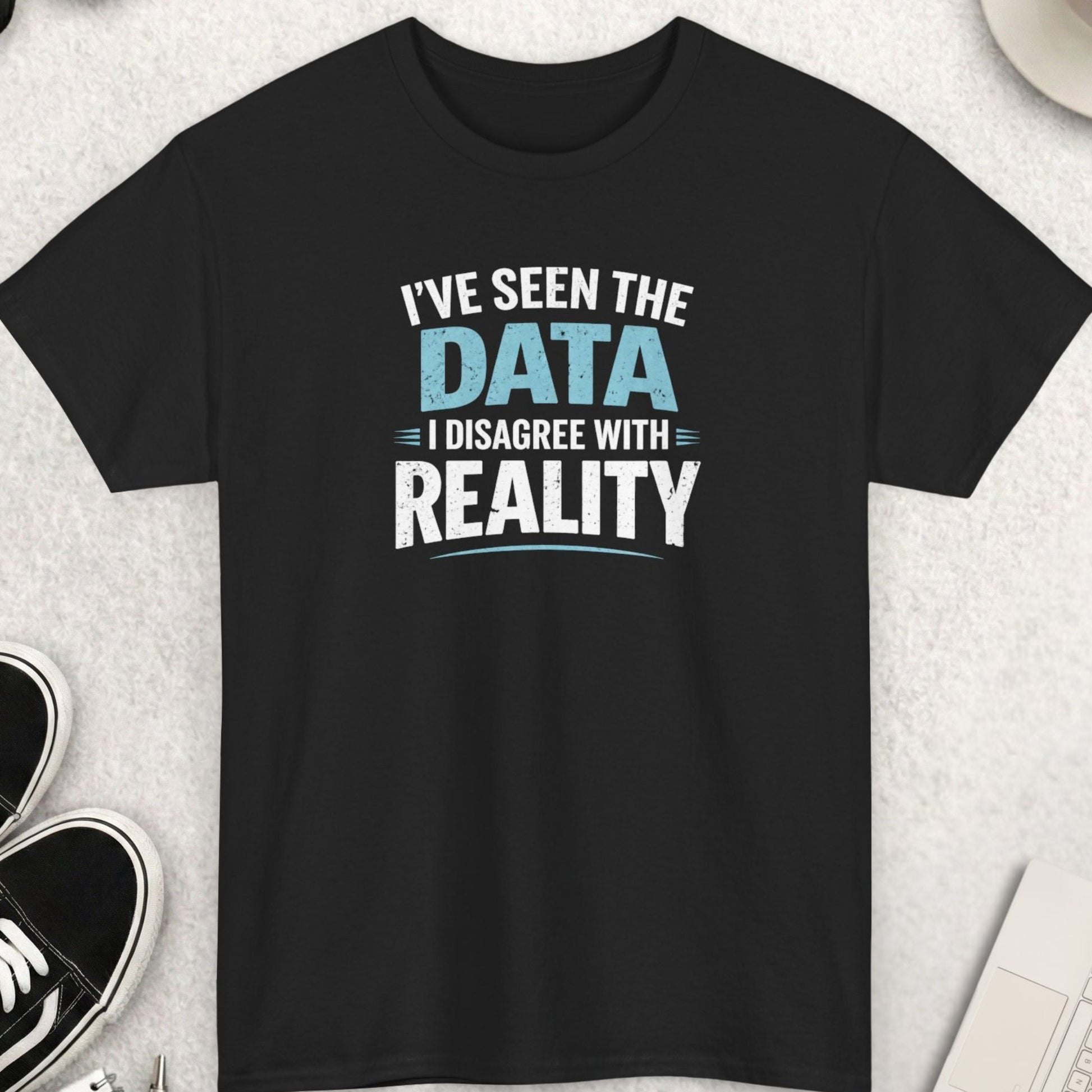 Black t-shirt with text 'I've seen the data, I disagree with reality' on a light gray surface with shoes, coffee, and sunglasses.