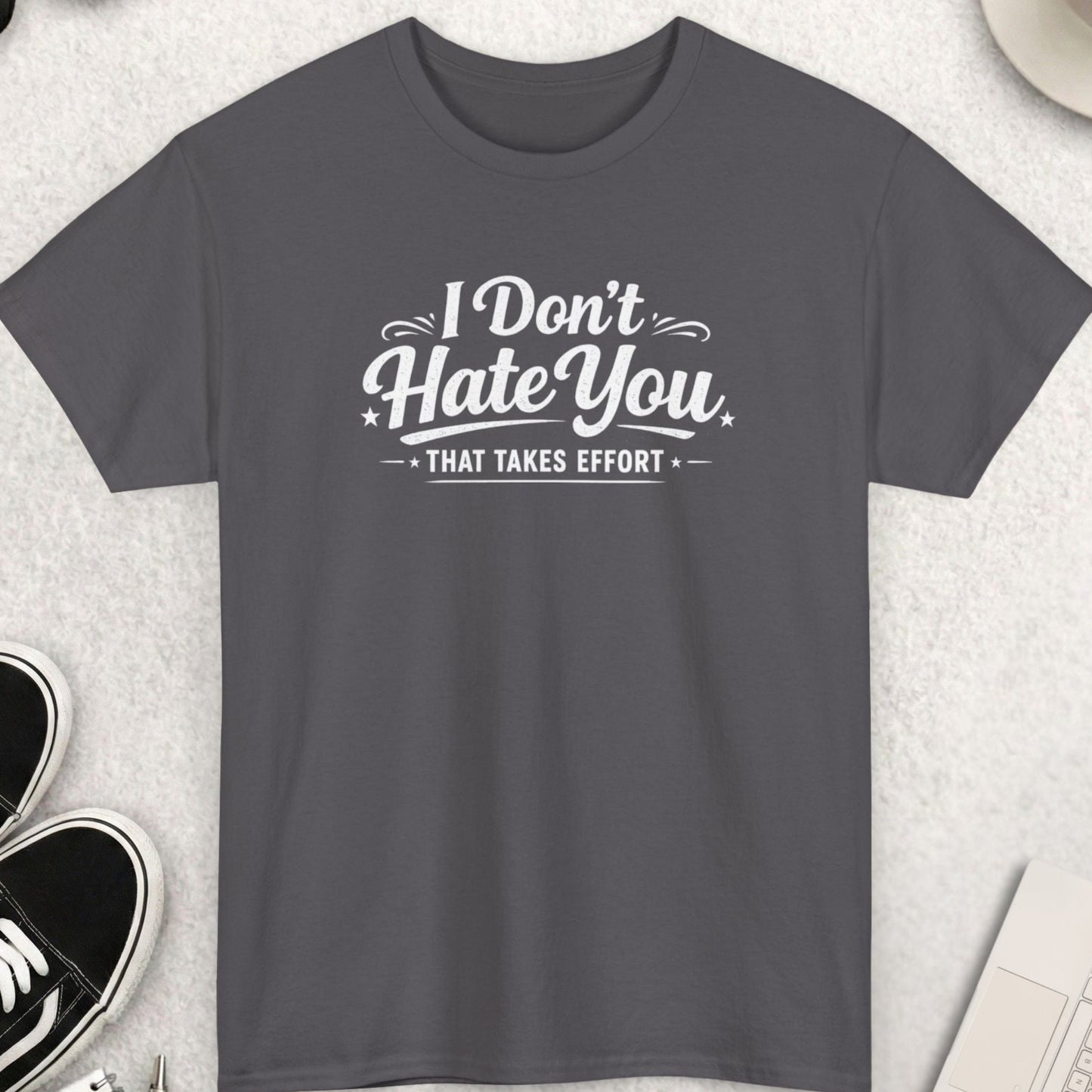 Gray t-shirt with 'I Don't Hate You That Takes Effort' text on a light gray surface with shoes, coffee, and accessories.
