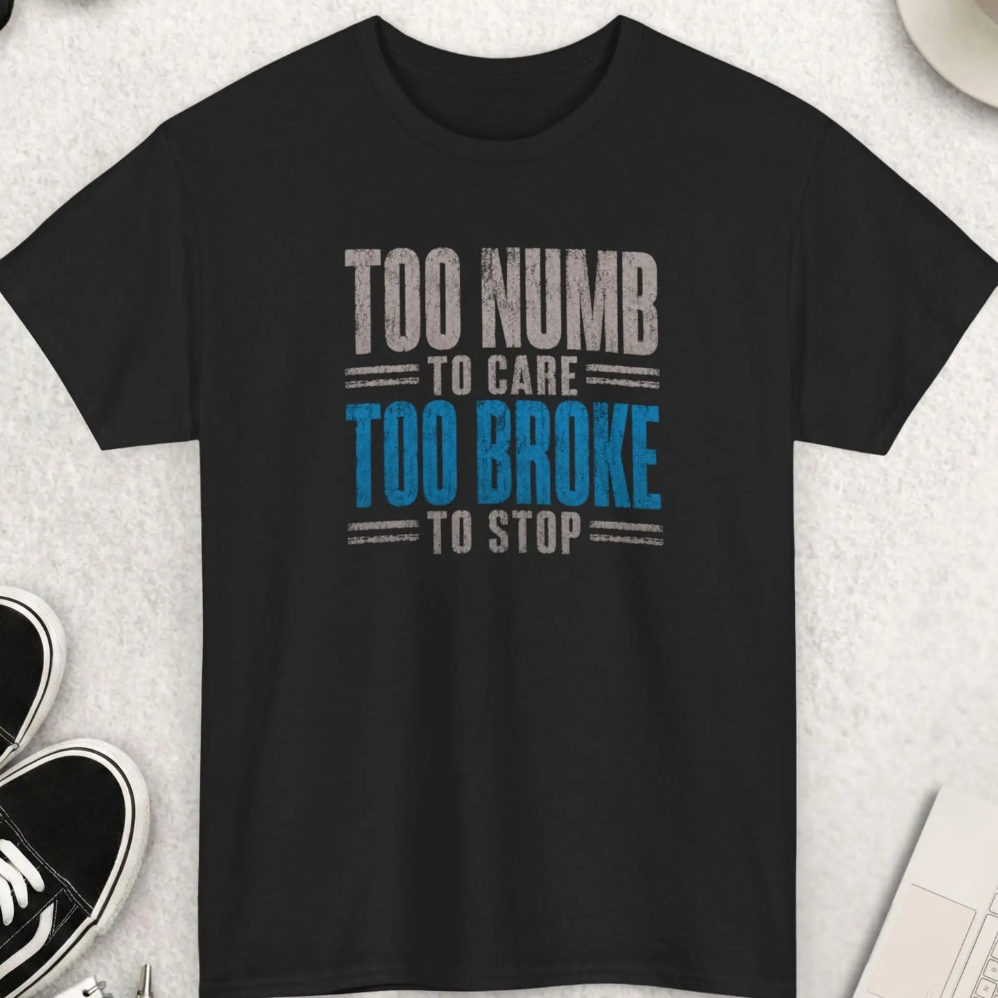 Black distressed typography graphic t-shirt featuring the phrase “Too Numb To Care Too Broke To Stop” in bold vintage cream and worn red lettering with a dark humor streetwear aesthetic by Mayhem Haus.