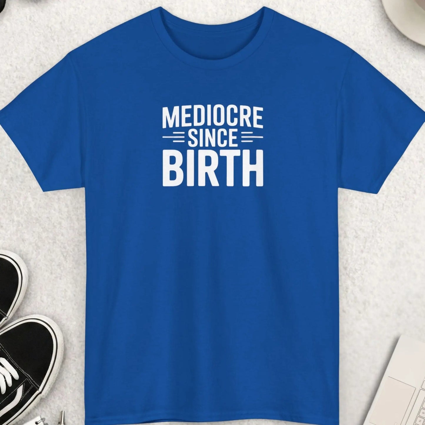 Blue t-shirt with 'Mediocre Since Birth' text on a light gray surface with shoes, sunglasses, and a cup.
