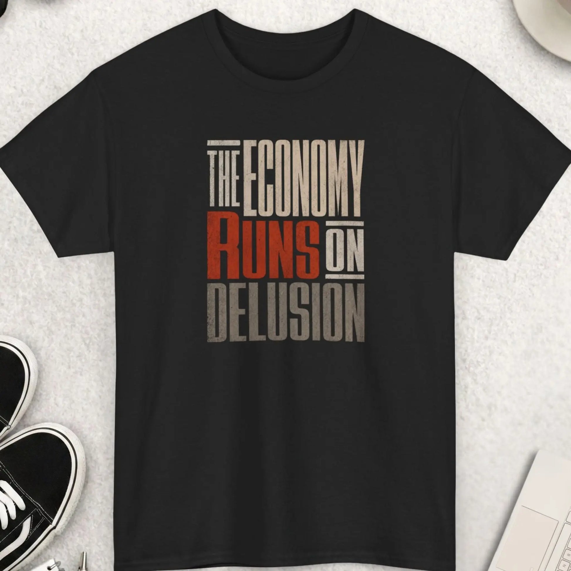 Distressed oversized typography t-shirt reading “The Economy Runs On Delusion” in cream, red, and gray lettering on a black shirt with dark humor economic satire streetwear aesthetic.