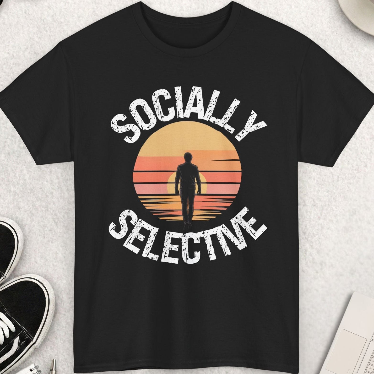 Black t-shirt with 'Socially Selective' design on a light gray surface with shoes, sunglasses, and coffee.