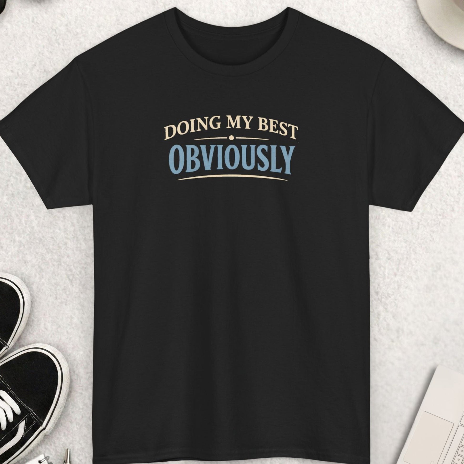 Black t-shirt with 'Doing my best Obviously' text on a light gray surface with shoes, coffee, and sunglasses.