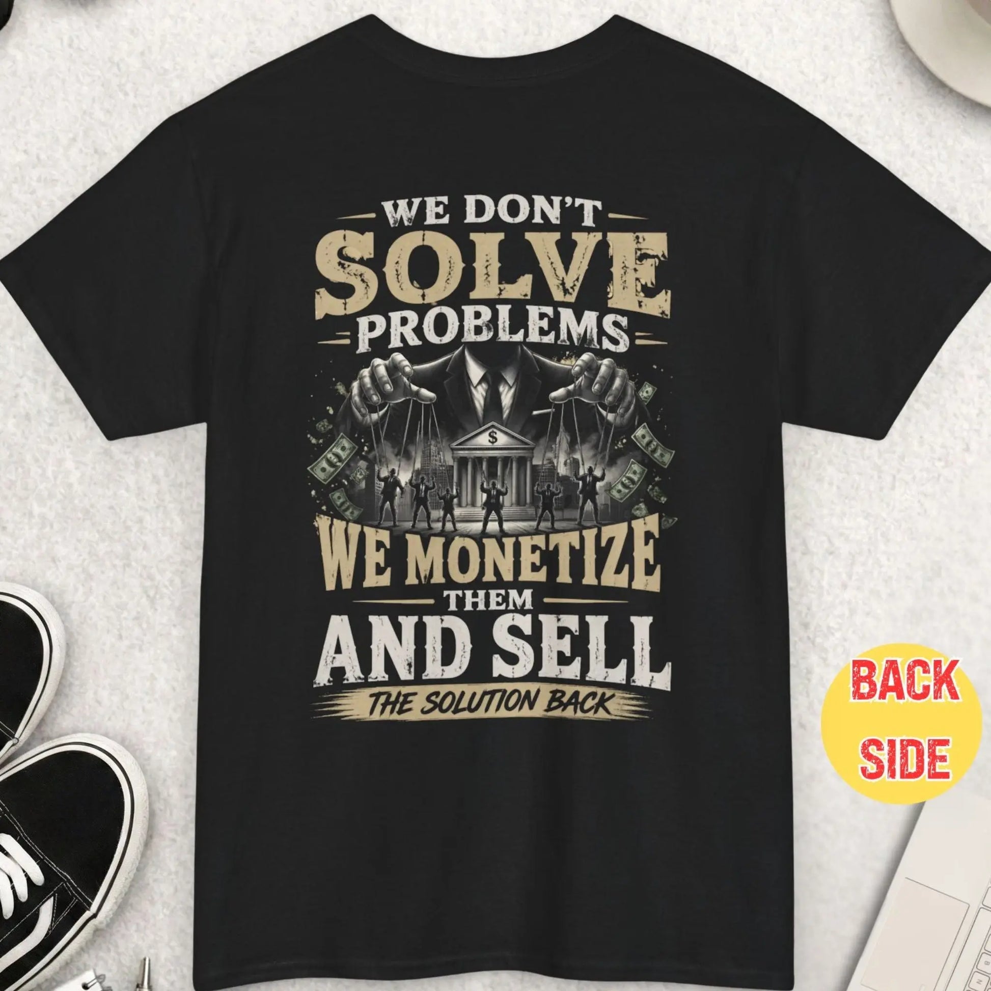 Black t-shirt with “we monetize problems and sell the solution back” message featuring puppet master controlling people and financial system imagery