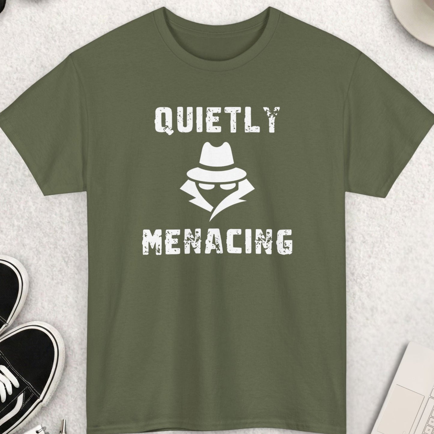 Green t-shirt with 'Quietly Menacing' design on a light gray surface with accessories.