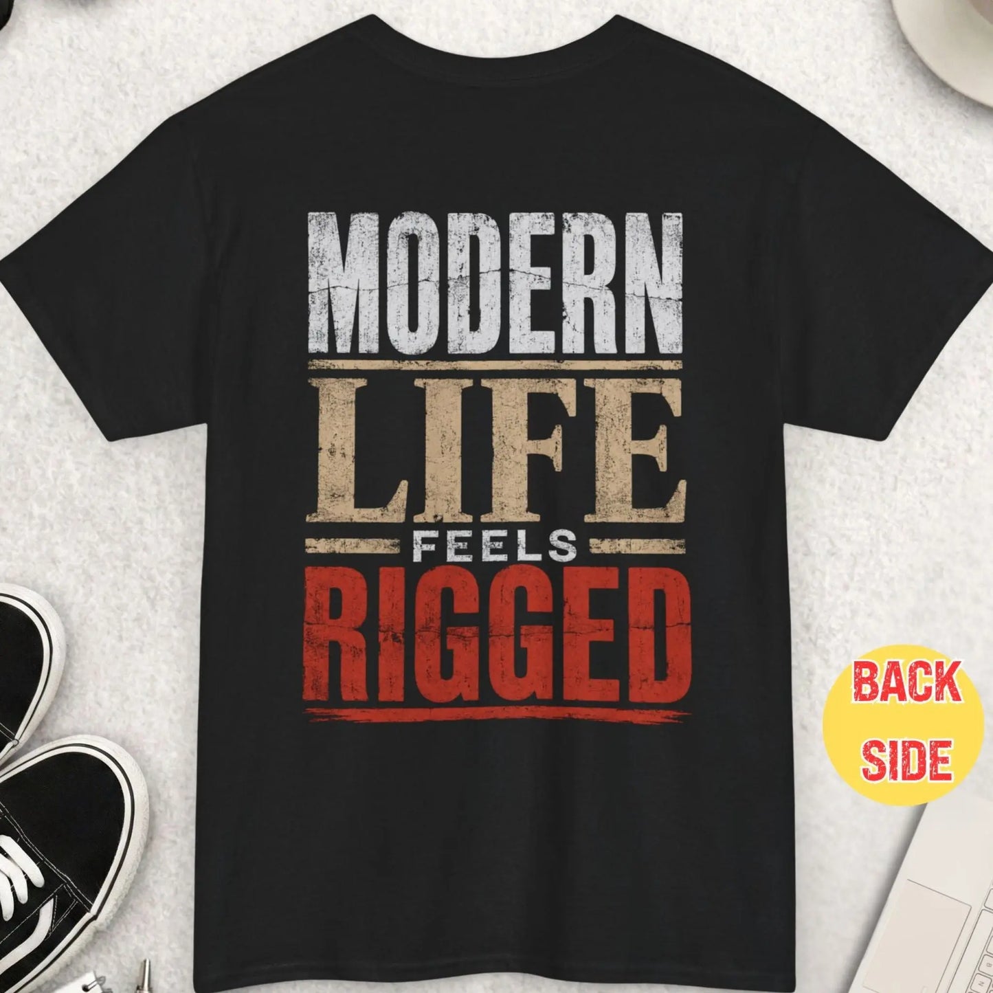 Black t-shirt featuring distressed bold typography reading “Modern Life Feels Rigged” in a high-contrast minimalist statement design with red accent lettering.