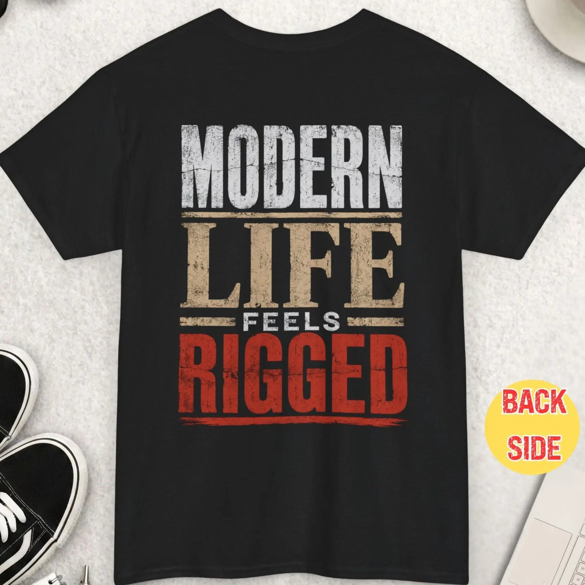 Black t-shirt featuring distressed bold typography reading “Modern Life Feels Rigged” in a high-contrast minimalist statement design with red accent lettering.