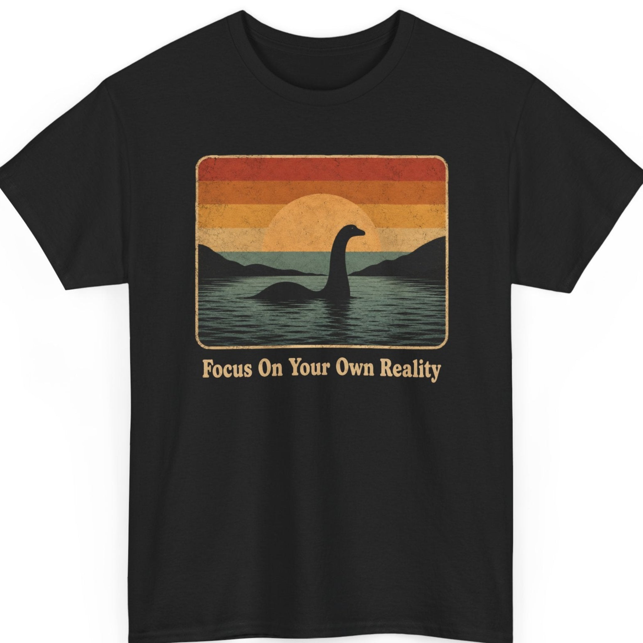 Mayhem Haus Loch Ness Monster graphic t-shirt on a black shirt featuring a vintage retro sunset over a lake with the silhouette of the Loch Ness Monster rising from the water above the phrase "Focus On Your Own Reality."