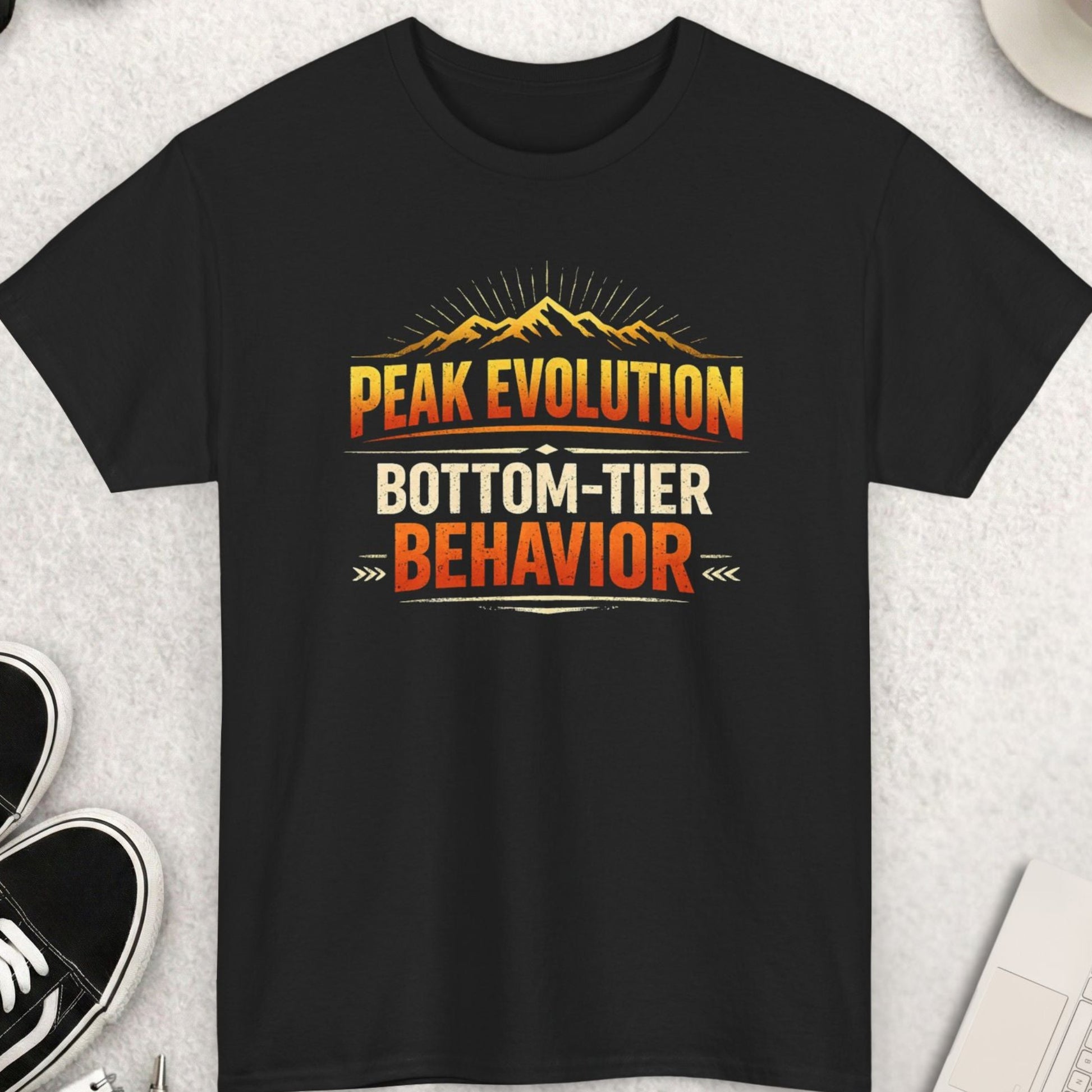 Black t-shirt with 'Peak Evolution Bottom-Tier Behavior' design on a light gray surface with shoes, coffee, and accessories.