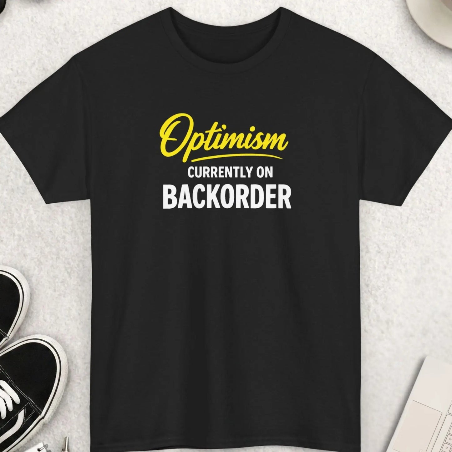 Black t-shirt with 'Optimism Currently on Back Order' text on a light gray surface with shoes, sunglasses, and a cup.