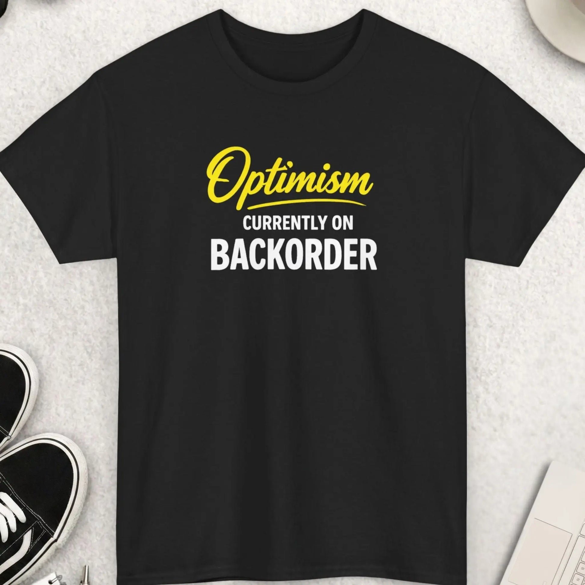 Black t-shirt with 'Optimism Currently on Back Order' text on a light gray surface with shoes, sunglasses, and a cup.
