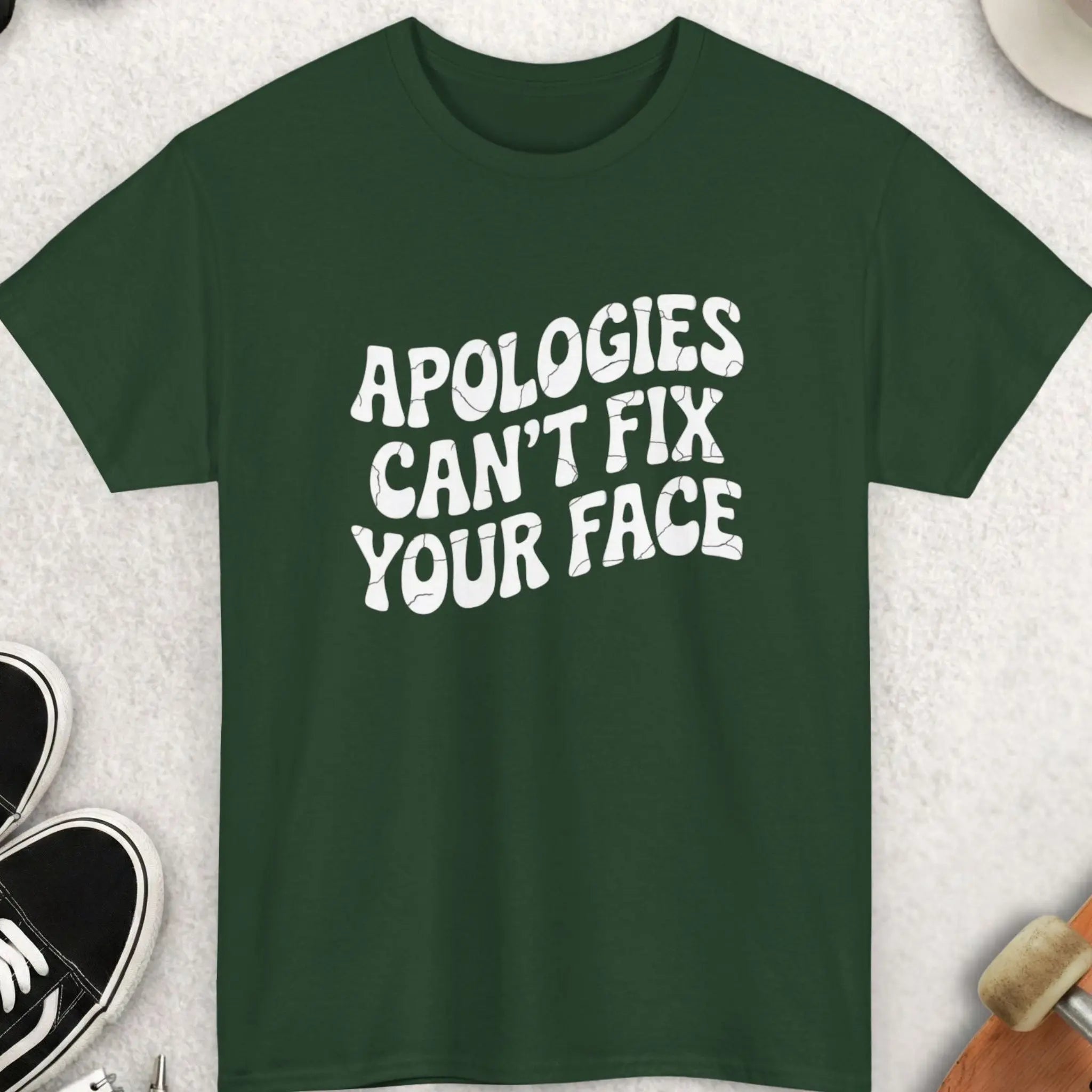 Green t-shirt with 'Apologies can't fix your face' text on a light gray surface with accessories.