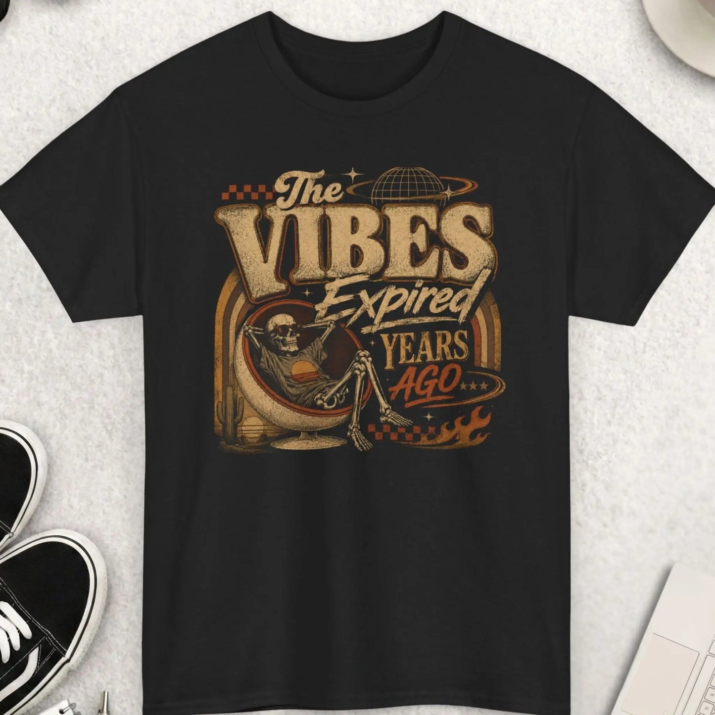 Black retro graphic t-shirt featuring the phrase “The Vibes Expired Years Ago” with distressed vintage typography and artwork of a relaxed skeleton in a retro chair surrounded by desert and cosmic imagery.