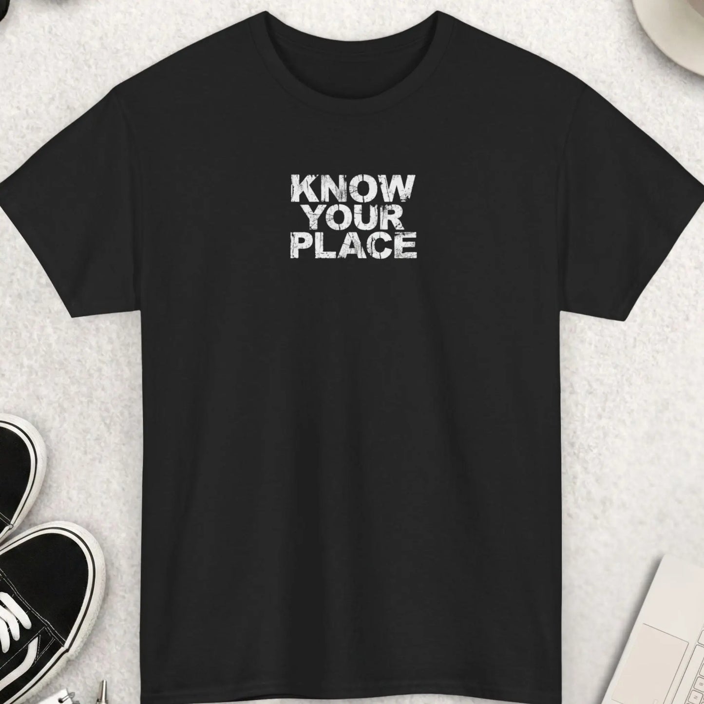 Black t-shirt with 'KNOW YOUR PLACE' text on a light gray surface with shoes, coffee, and accessories.
