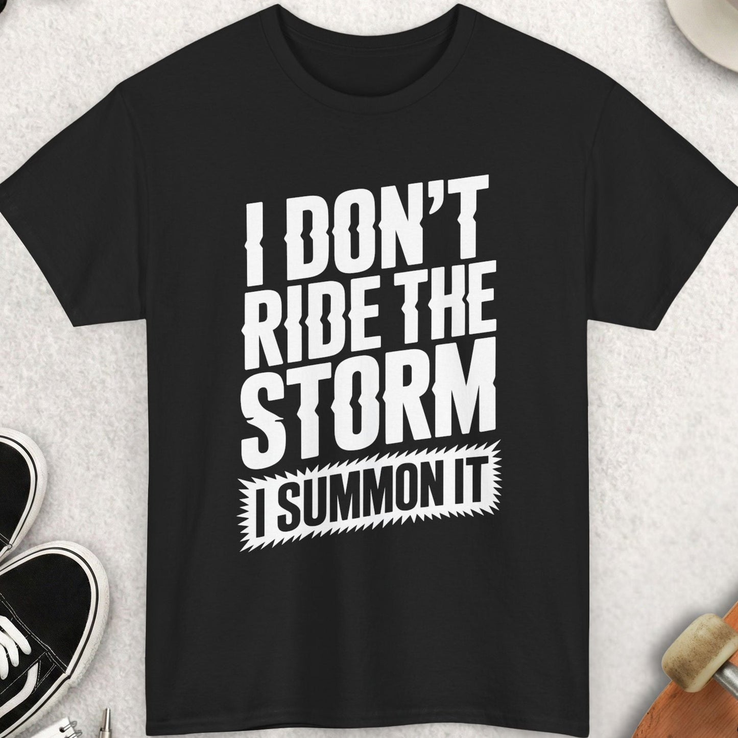 Black t-shirt with 'I don't ride the storm, I summon it' text on a light gray surface with shoes, sunglasses, and a cup.