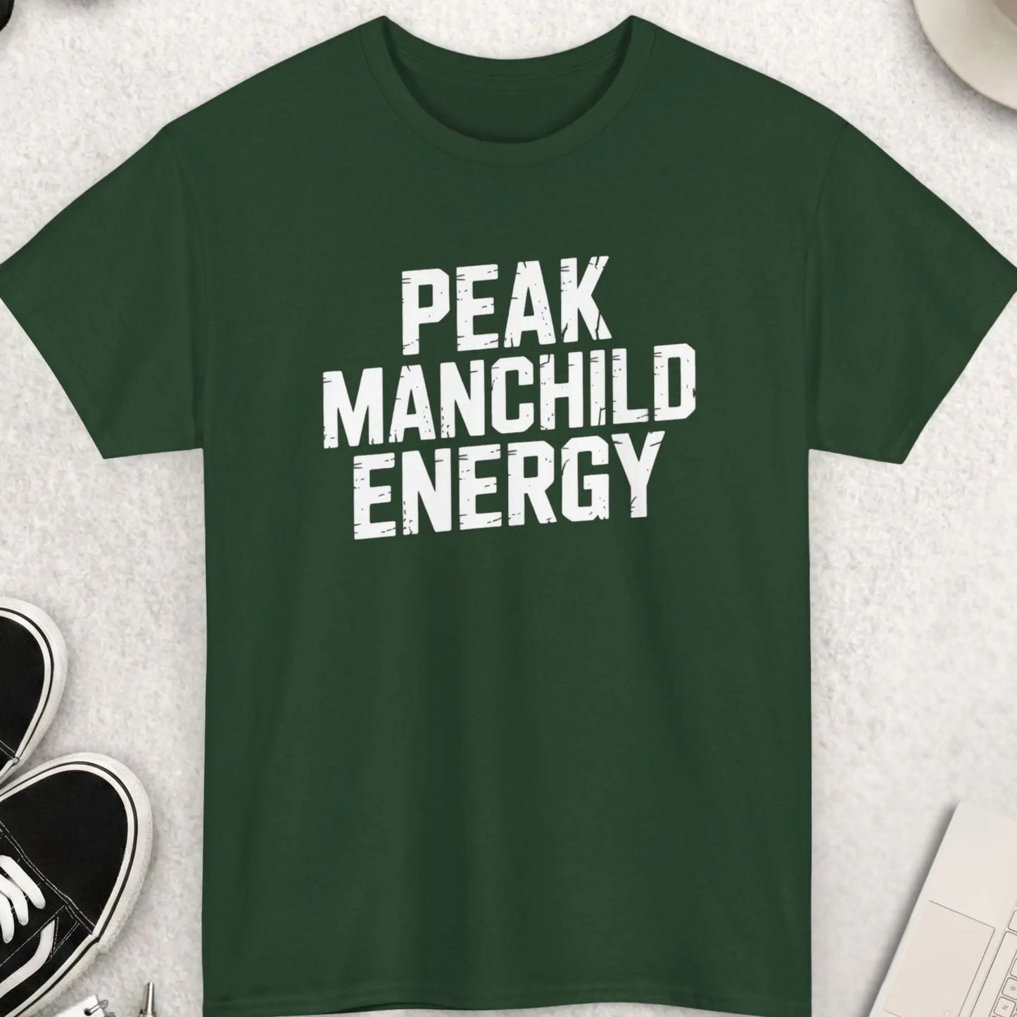 Green t-shirt with 'PEAK MANCHILD ENERGY' text on a light gray surface with accessories.