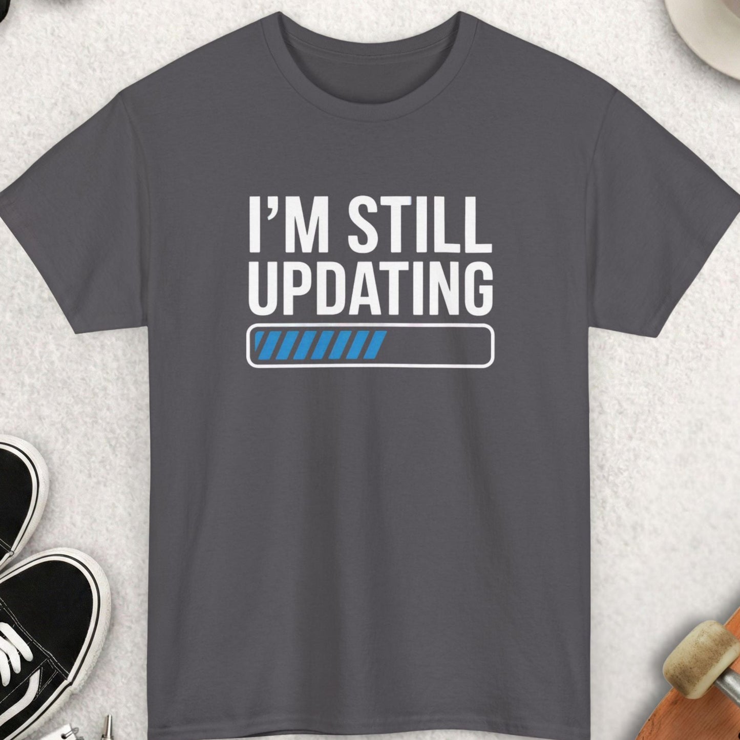 Gray t-shirt with 'I'm Still Updating' text on a light gray surface with accessories.