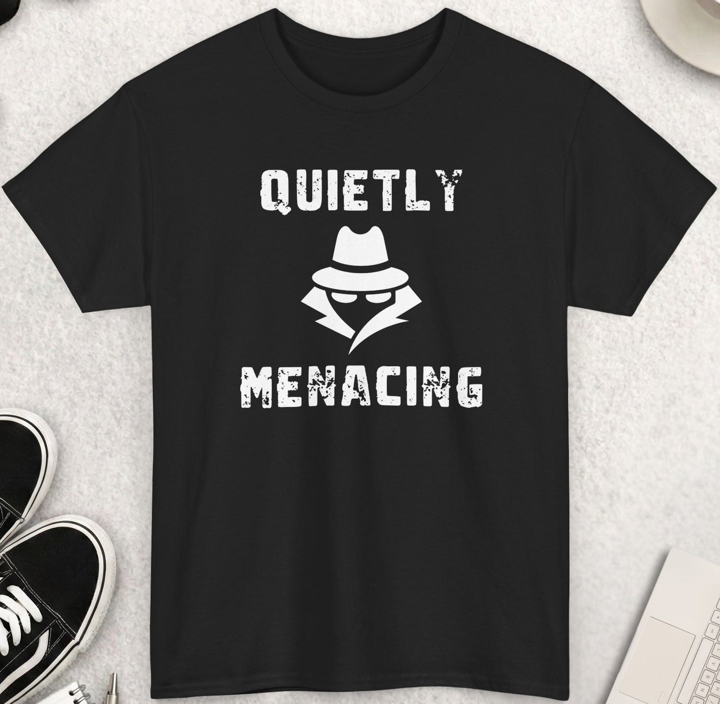 Black t-shirt with 'Quietly Menacing' design on a light gray surface with shoes, sunglasses, and a cup of coffee.