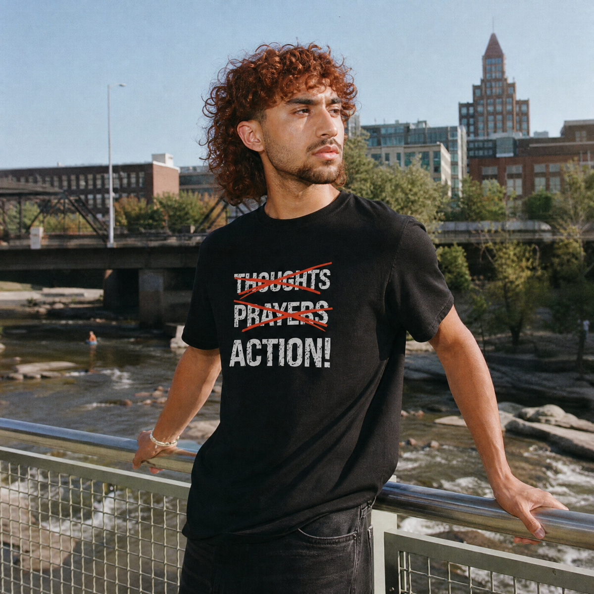 Mayhem Haus Thoughts Prayers Action T-Shirt on a person with curly hair, standing by a river